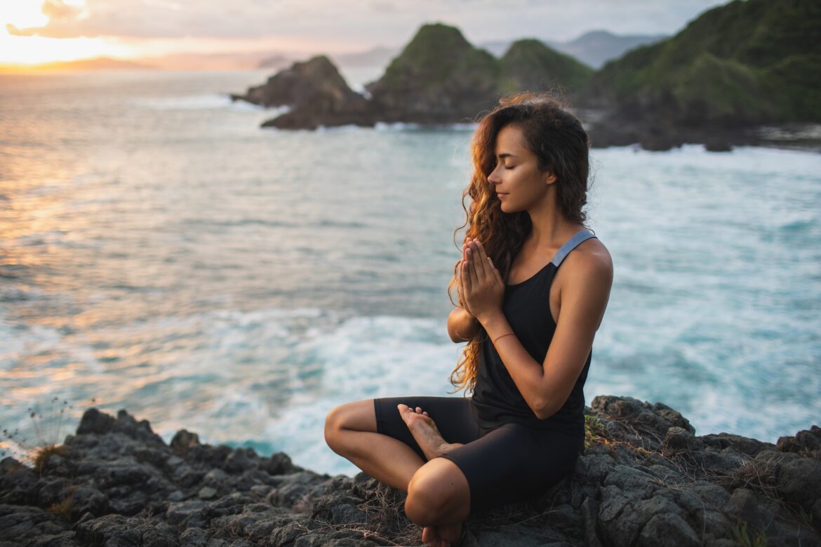 Young woman praying and meditating alone at sunset with beautiful ocean and mountain view.